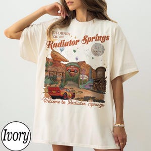 K&ouml;nnte beinhalten: Elfenbeinfarbenes T-Shirt mit einem Radiator Springs-Motiv, einschlie&szlig;lich des Textes "LIFORNIA, Est. 2017" und "Welcome to Radiator Springs." Das Design zeigt ein rotes Rennauto und eine W&uuml;stenlandschaft.