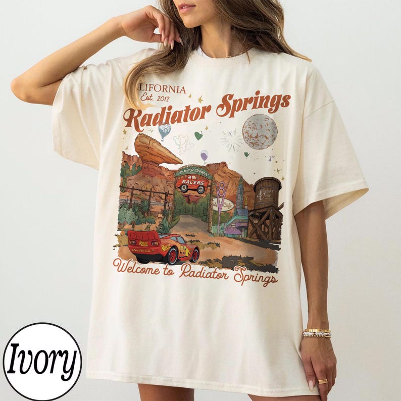 K&ouml;nnte beinhalten: Elfenbeinfarbenes T-Shirt mit einem Radiator Springs-Motiv, einschlie&szlig;lich des Textes "LIFORNIA, Est. 2017" und "Welcome to Radiator Springs." Das Design zeigt ein rotes Rennauto und eine W&uuml;stenlandschaft.