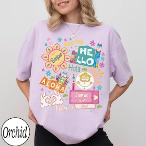 Może przedstawiać: Jasnofioletowy t-shirt z kr&oacute;tkim rękawem z kolorowym nadrukiem graficznym przedstawiającym r&oacute;żne międzynarodowe powitania, w tym Bonjour, Hello, Hola, Aloha, Jambo i Hej, wraz ze stylizowanymi ilustracjami kwiat&oacute;w, zamku i dłoni.