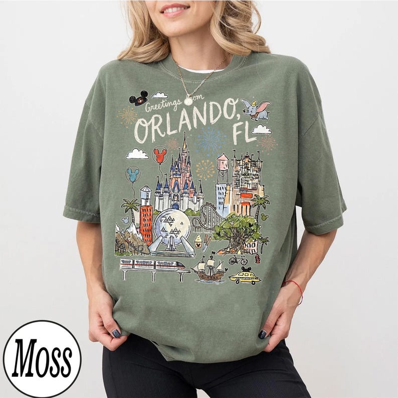 May include: Olive green t-shirt with a detailed illustration of Orlando, Florida landmarks. The design includes a castle, Spaceship Earth, and the text "Greetings from Orlando, FL". The shirt has a relaxed fit.