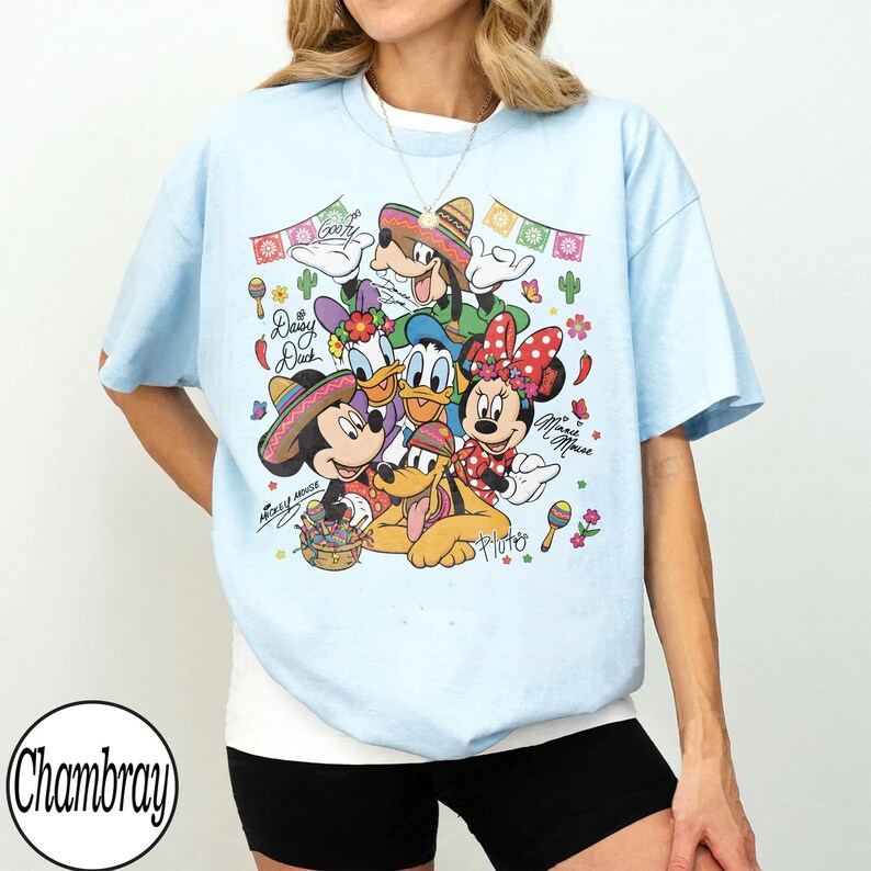 May include: Pale blue t-shirt with a vibrant graphic of Disney characters Mickey Mouse, Minnie Mouse, Donald Duck, Daisy Duck, Goofy, and Pluto in Mexican-themed outfits. The design includes sombreros, maracas, and the text "Mickey Mouse House".