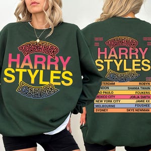 Pu&ograve; includere: Una felpa girocollo verde scuro con una stampa grafica. La parte anteriore mostra il testo HARRY STYLES TOGETHER in rosa e giallo. Il retro presenta lo stesso testo con un elenco di citt&agrave; e nomi tra cui Amsterdam, London e Shania Twain.