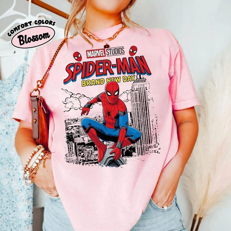 Może przedstawiać: R&oacute;żowa koszulka z grafiką Spider-Mana z Marvel Studios. Projekt zawiera postać Spider-Mana w czerwono-niebieskim kostiumie, napis "Spider-Man Brand New Day" i logo Marvel Studios.