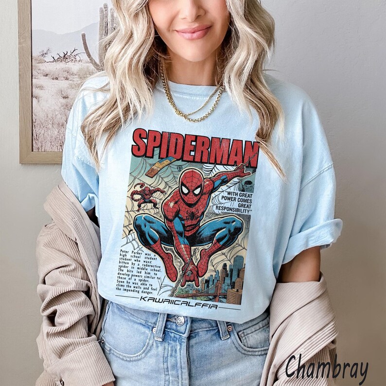May include: Light blue t-shirt featuring a vintage comic book style graphic of Spider-Man. The graphic includes the word "SPIDERMAN" in red, and the phrase "WITH GREAT POWER COMES GREAT RESPONSIBILITY". The shirt has rolled-up sleeves.