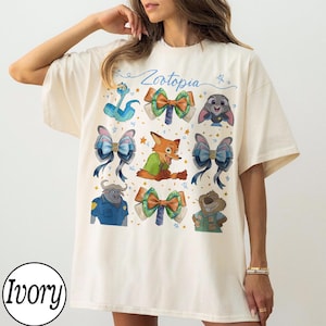 May include: Ivory-colored t-shirt featuring colorful illustrations of characters and bows from the movie "Zootopia." The design includes a fox, bunny, and other characters. The word "Zootopia" is written in cursive.