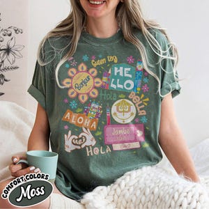 Może przedstawiać: Mchowo-zielony t-shirt z kr&oacute;tkim rękawem z kolorowym nadrukiem graficznym zawierającym r&oacute;żne międzynarodowe powitania, takie jak Bonjour, Hello, Hola, Aloha, Jambo i Hej. Projekt zawiera stylizowane kwiaty, bilety oraz dłonie z kresk&oacute;wek tworzące kształt serca, wszystko w postarzanym stylu.