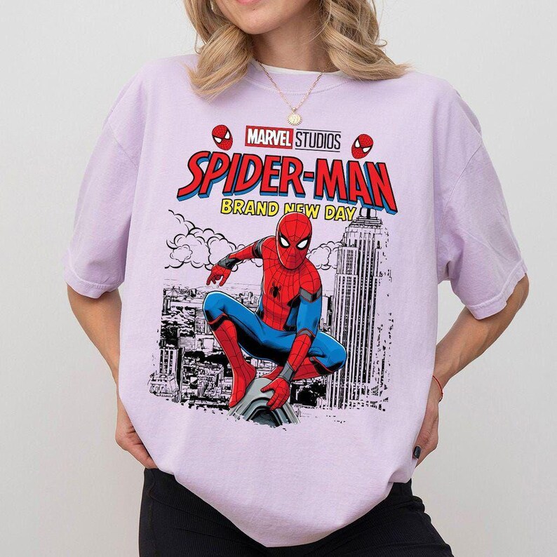 Może przedstawiać: Jasnofioletowa koszulka z grafiką Spider-Mana w pozycji kucznej na tle panoramy miasta. Logo Marvel Studios i tekst "Spider-Man Brand New Day" są wyświetlane nad postacią.