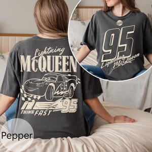 May include: Dark gray t-shirt featuring Lightning McQueen graphics. The front displays a large "95" with "Lightning McQueen" text. The back showcases a car illustration, the number "95", and the words "Lightning McQueen" and "Think Fast".