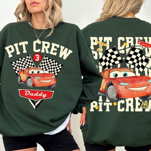 May include: Dark green sweatshirt with a graphic of Lightning McQueen from the movie Cars. The front features the text "PIT CREW" above a checkered flag and the number 3. Below is the word "Daddy" in a red and white design.