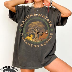 May include: Dark gray t-shirt featuring a circular design with the words "HAKUNA MATATA" and "IT MEANS NO WORRIES". The design includes a tree, Simba, Timon, and Pumbaa. The shirt also has the words "COMFORT COLORS" and "Pepper".
