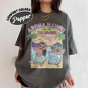 May include: Dark grey t-shirt with a colourful graphic. The graphic reads "Aloha Hawaii Come Visit The Islands" and depicts two cartoon characters in sunglasses and hula skirts on a beach with palm trees.