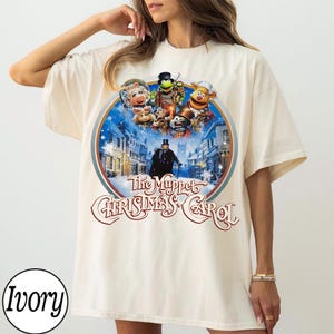 Puede incluir: Una camiseta de manga corta color marfil con un gráfico circular de The Muppet Christmas Carol. El estampado muestra varios personajes de los Muppets sobre una escena callejera nevada con una figura central con sombrero de copa y el texto The Muppet Christmas Carol.