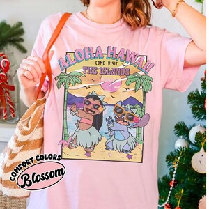 May include: Pale pink t-shirt with a graphic of two cartoon characters on a beach, and the text "Aloha Hawaii! Come Visit The Islands." The shirt also has the words "Comfort Colors" and "Blossom" printed on it.