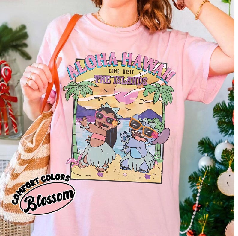 May include: Pale pink t-shirt with a graphic of two cartoon characters on a beach, and the text "Aloha Hawaii! Come Visit The Islands." The shirt also has the words "Comfort Colors" and "Blossom" printed on it.