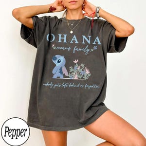 May include: Dark gray t-shirt featuring the text "OHANA means family" in blue, along with a cartoon character and floral accents. The shirt also includes the phrase "nobody gets left behind or forgotten".
