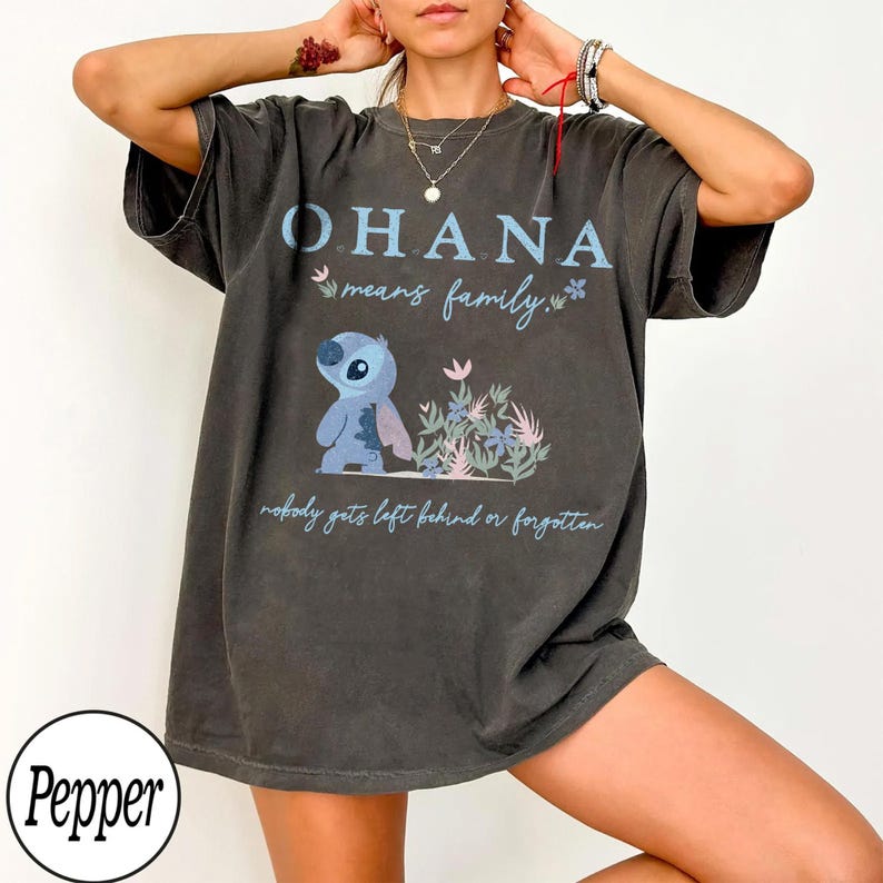 May include: Dark gray t-shirt featuring the text "OHANA means family" in blue, along with a cartoon character and floral accents. The shirt also includes the phrase "nobody gets left behind or forgotten".
