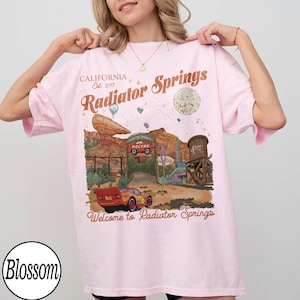 K&ouml;nnte beinhalten: Hellrosa T-Shirt mit einem Radiator Springs-Motiv, mit Text wie "CALIFORNIA Est. 2017" und "Radiator Springs". Das Design zeigt ein Cartoon-Auto, eine W&uuml;stenlandschaft und die Worte "Welcome to Radiator Springs".