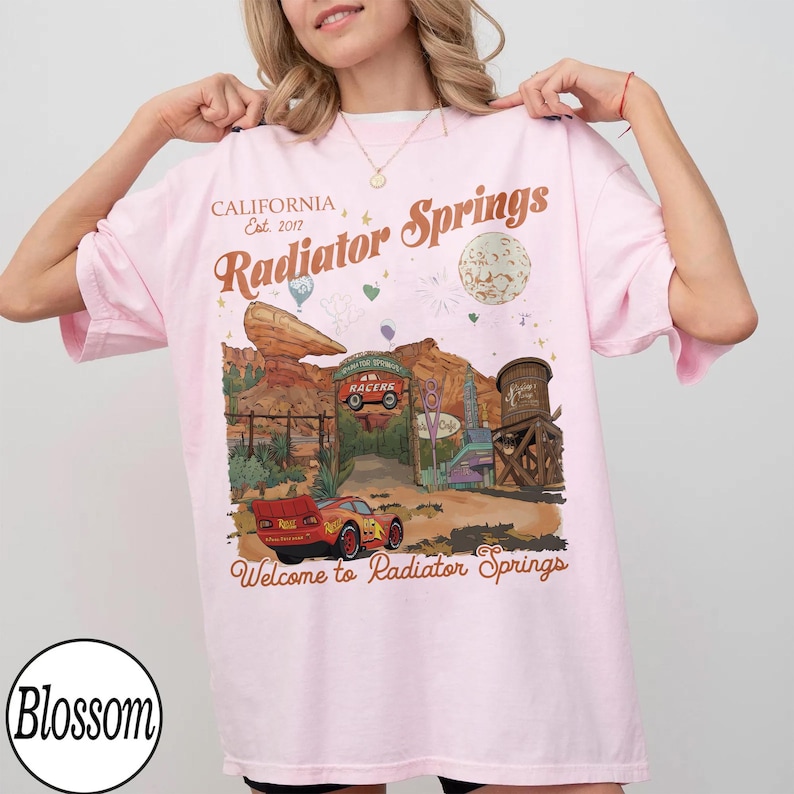 K&ouml;nnte beinhalten: Hellrosa T-Shirt mit einem Radiator Springs-Motiv, mit Text wie "CALIFORNIA Est. 2017" und "Radiator Springs". Das Design zeigt ein Cartoon-Auto, eine W&uuml;stenlandschaft und die Worte "Welcome to Radiator Springs".
