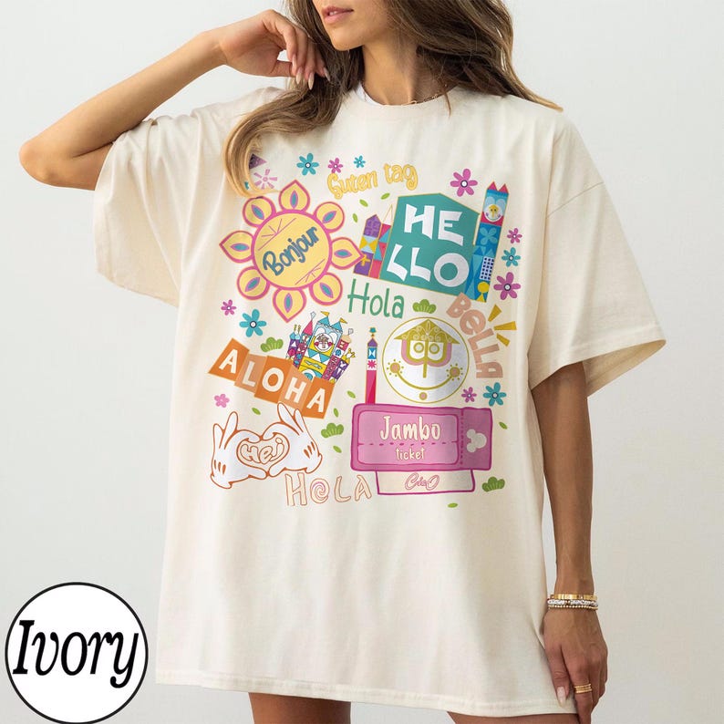Może przedstawiać: T-shirt z kr&oacute;tkim rękawem w kolorze kości słoniowej z kolorowym nadrukiem graficznym przedstawiającym r&oacute;żne międzynarodowe powitania, takie jak Bonjour, Hello, Hola, Bella, Aloha, Jambo i Hej. Wz&oacute;r zawiera stylizowane kwiaty, bilet i dłonie z kresk&oacute;wek.