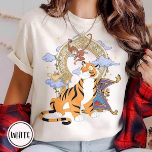May include: White t-shirt featuring a cartoon tiger with black stripes, a monkey, and a magic carpet. The design includes a gold-colored archway, clouds, and a magic lamp. The word "WHITE" is in a white circle at the bottom.