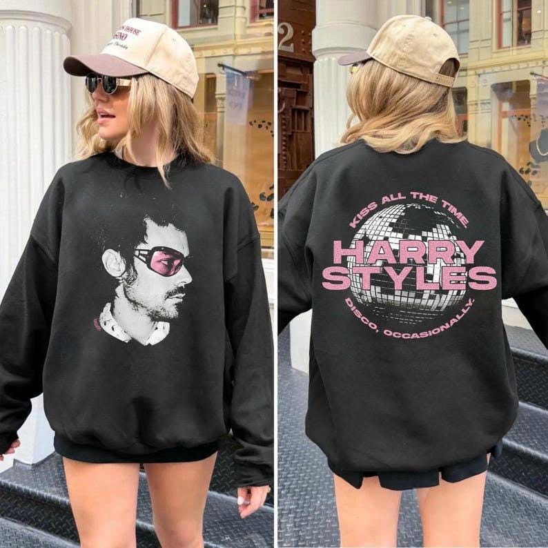 May include: Black sweatshirt featuring a side profile portrait of a man wearing pink sunglasses. The back of the sweatshirt has the text "KISS ALL THE TIME" and "HARRY STYLES DISCO, OCCASIONALLY" in pink and white.
