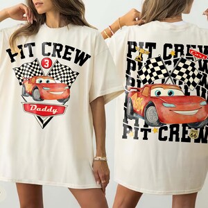 May include: Ivory-colored t-shirt featuring a graphic of a red race car with the text "PIT CREW" and "Daddy". The design includes checkered flags and the number 3. The back of the shirt also has the same design.