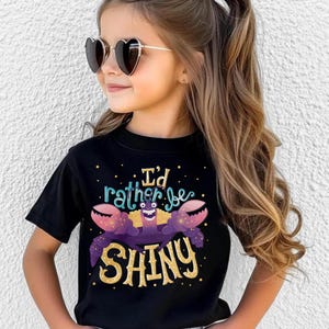 Peut inclure: T-shirt noir avec l'inscription "I'd rather be SHINY" et un dessin de crabe. Le crabe est violet avec des détails dorés. La personne porte des lunettes de soleil en forme de cœur.