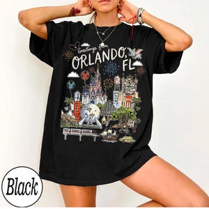 May include: Black t-shirt with a colorful graphic of Orlando, Florida landmarks, including a castle, fireworks, and the text "Greetings from Orlando, FL." The shirt has a relaxed fit.