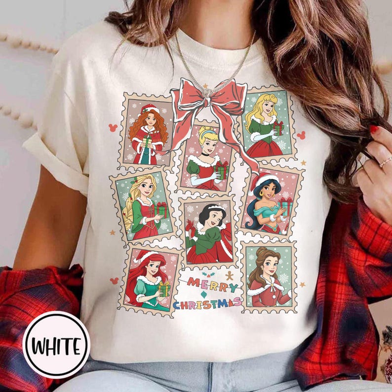 May include: White t-shirt featuring a festive design with various Disney princesses in Christmas-themed postage stamp frames. A large red bow and the words "MERRY CHRISTMAS" are also present. The shirt has short sleeves.