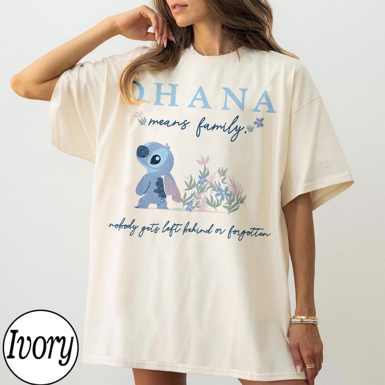 May include: Ivory-colored t-shirt featuring the word "OHANA" in light blue, with the phrase "means family" and a cartoon character. The shirt also has the text "nobody gets left behind or forgotten."