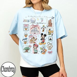 K&ouml;nnte beinhalten: Hellblaues T-Shirt mit einem Disney-Motiv. Das Design zeigt Figuren wie Mickey Mouse und Donald Duck, sowie den Text "The Most Magical Place on Earth" und das Jahr "2026".