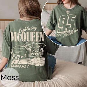 May include: Moss green t-shirt featuring Lightning McQueen graphics. The front displays "95" and "Lightning McQueen" text. The back showcases a race car illustration with "Lightning McQueen", "95", and "Think Fast" text. A comfortable, casual tee.