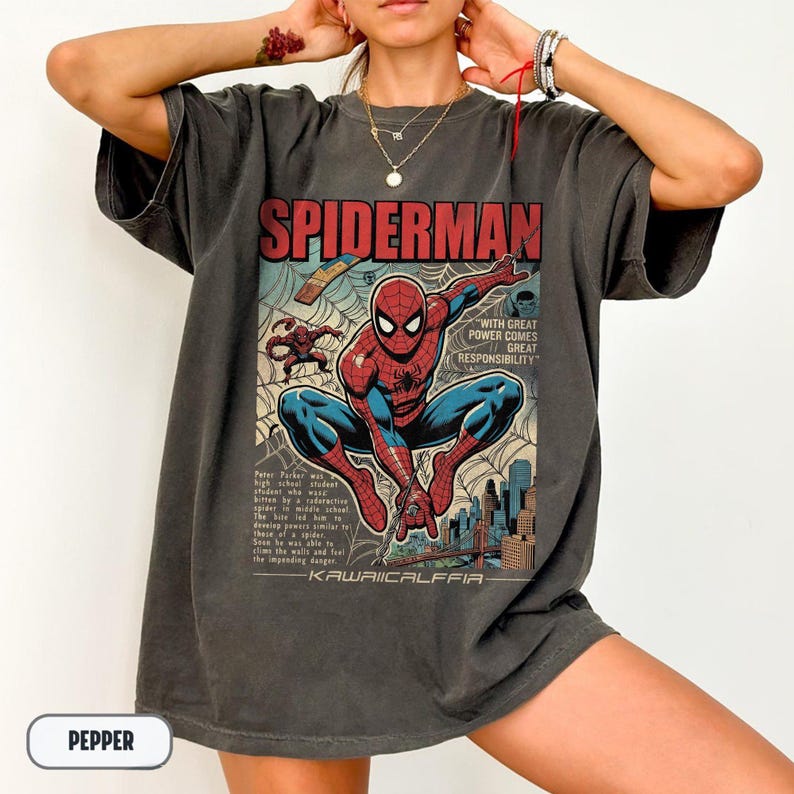 May include: Dark gray t-shirt featuring a comic book style graphic of Spider-Man. The design includes the word "SPIDERMAN" in red, the phrase "WITH GREAT POWER COMES GREAT RESPONSIBILITY", and a cityscape background. The shirt also has the word "PEPPER" on the bottom.