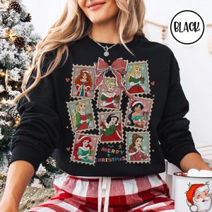 May include: Black sweatshirt featuring a festive design with princess illustrations in postage stamp frames. A large pink bow and the words "Merry Christmas" are also featured. The sweatshirt has a crew neck and long sleeves.