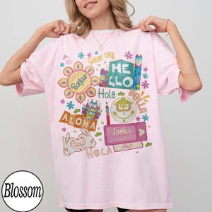 Może przedstawiać: Jasnor&oacute;żowy t-shirt z kolorowym nadrukiem graficznym zawierającym r&oacute;żne międzynarodowe powitania, w tym Bonjour, Guten Tag, Hello, Hola, Bella, Aloha, Jambo i Hej. Wz&oacute;r zawiera stylizowane ikony zamk&oacute;w, kwiaty oraz dłonie z kresk&oacute;wek.