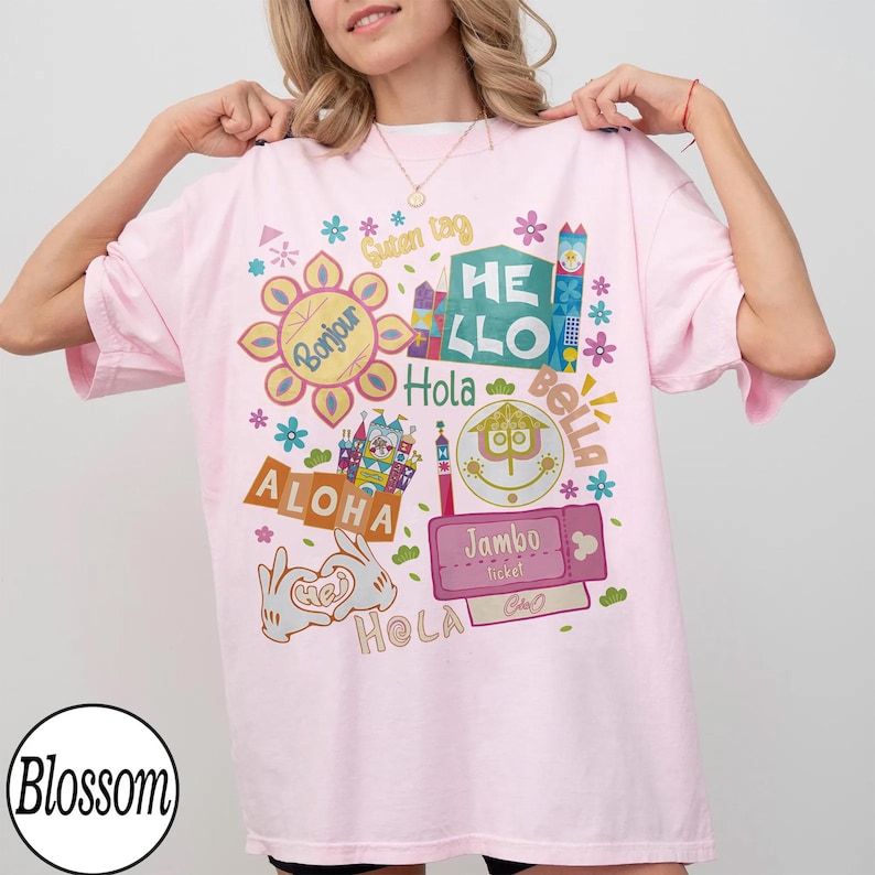Może przedstawiać: Jasnor&oacute;żowy t-shirt z kolorowym nadrukiem graficznym zawierającym r&oacute;żne międzynarodowe powitania, w tym Bonjour, Guten Tag, Hello, Hola, Bella, Aloha, Jambo i Hej. Wz&oacute;r zawiera stylizowane ikony zamk&oacute;w, kwiaty oraz dłonie z kresk&oacute;wek.