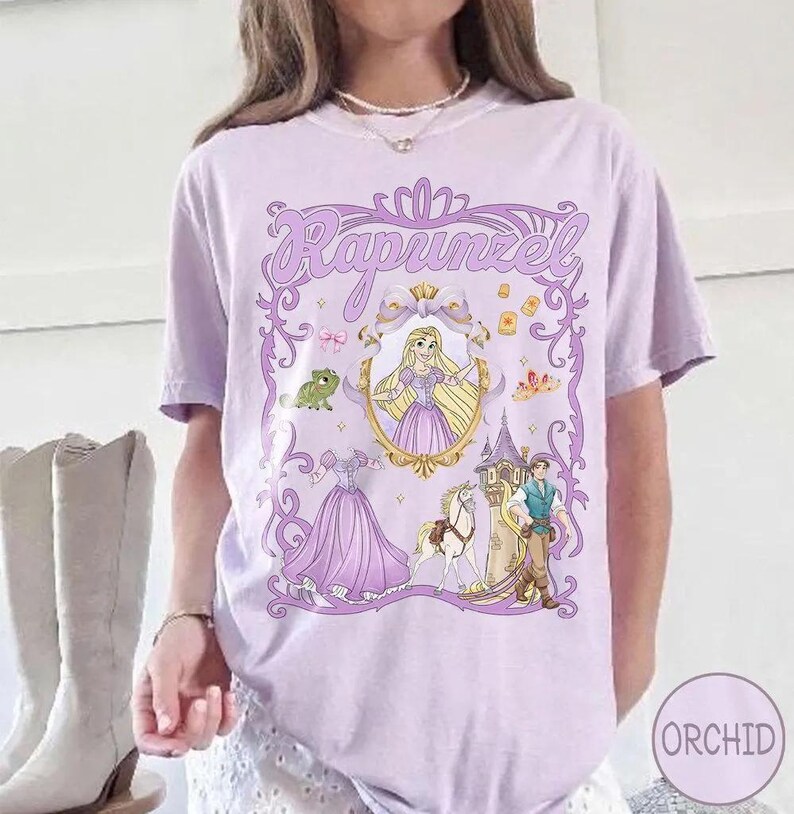 May include: A light purple t-shirt featuring a graphic of Rapunzel from the Disney movie Tangled. The design includes Rapunzel in a gold frame, a frog, a dress, a castle, and the text "Rapunzel" in a decorative font.