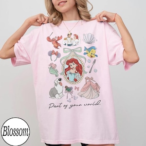 Puede incluir: Camiseta rosa claro con ilustraciones de dibujos animados, incluyendo personajes y un vestido. El texto "Part of your world" tambi&eacute;n est&aacute; presente.