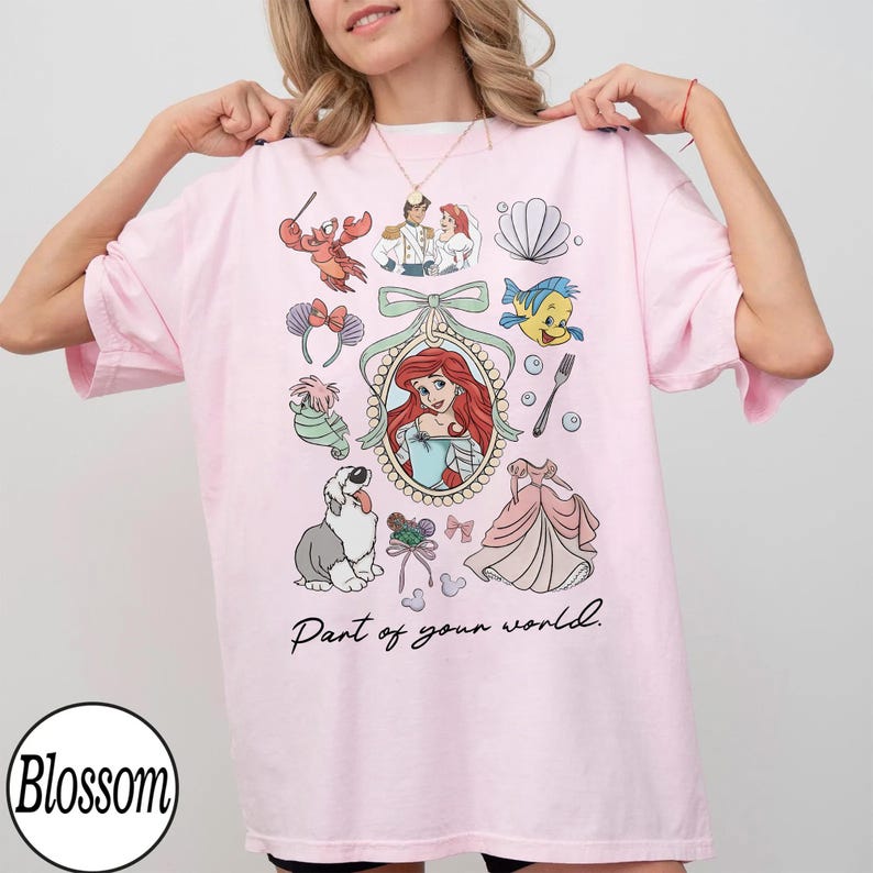 Puede incluir: Camiseta rosa claro con ilustraciones de dibujos animados, incluyendo personajes y un vestido. El texto "Part of your world" tambi&eacute;n est&aacute; presente.
