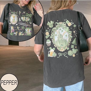 May include: Dark gray t-shirt with a front graphic of Tinkerbell and other fairy-themed images. The back of the shirt features a castle, shamrocks, and other green and gold graphics. The shirt has short sleeves and a crew neck.
