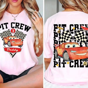 May include: Pink t-shirt with a graphic of a red race car and the text "PIT CREW Daddy" on the front. The back of the shirt features the text "PIT CREW" and a similar car graphic. Checkered flags and stars are also included in the design.