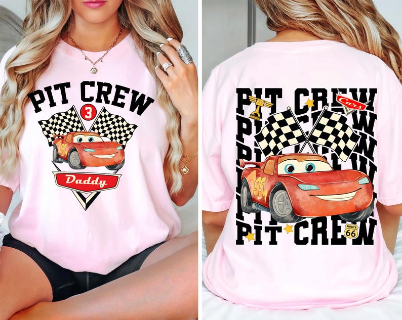 May include: Pink t-shirt with a graphic of a red race car and the text "PIT CREW Daddy" on the front. The back of the shirt features the text "PIT CREW" and a similar car graphic. Checkered flags and stars are also included in the design.