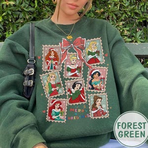 May include: Forest green sweatshirt featuring a design of various princesses in Christmas-themed postage stamp frames. A large red bow is above the stamps, and the words "Merry Christmas" are printed below. The sweatshirt is a solid forest green color.