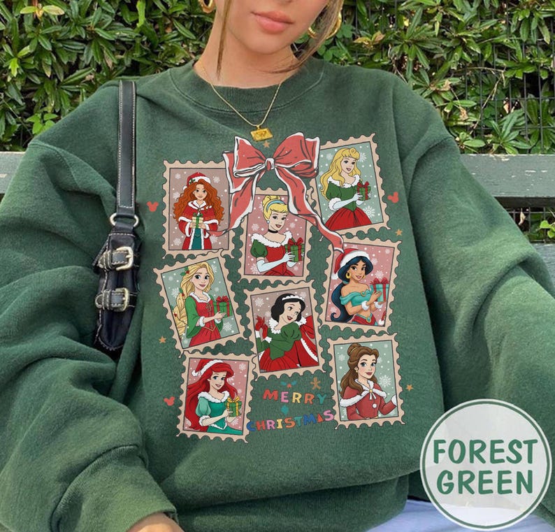 May include: Forest green sweatshirt featuring a design of various princesses in Christmas-themed postage stamp frames. A large red bow is above the stamps, and the words "Merry Christmas" are printed below. The sweatshirt is a solid forest green color.
