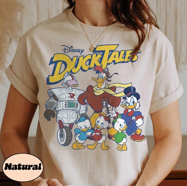 May include: A beige t-shirt featuring the Disney DuckTales logo in yellow and blue, with a graphic of the DuckTales characters. The word "Natural" is printed on the bottom left of the shirt.