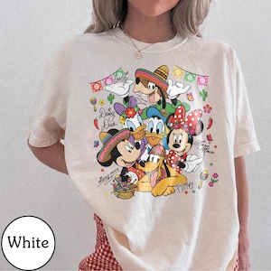 May include: Cream-coloured t-shirt with a vibrant graphic of Disney characters Mickey Mouse, Minnie Mouse, Donald Duck, Daisy Duck, Goofy, and Pluto, wearing sombreros. The design includes text and decorative elements.