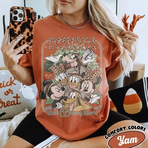 May include: Burnt orange t-shirt featuring a graphic of Disney characters, including Mickey and Minnie Mouse, Donald and Daisy Duck, and Goofy. The shirt has the text "ANIMAL KINGDOM" and "Walt Disney World". The shirt is from Comfort Colors.
