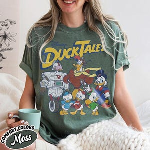 May include: Green t-shirt featuring the Disney DuckTales logo and cartoon characters. The design includes Scrooge McDuck, Huey, Dewey, Louie, and other characters. The shirt is a comfort colors moss color.