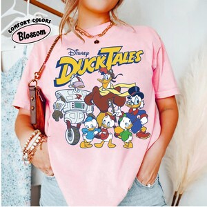 May include: Pink t-shirt featuring the Disney DuckTales logo and characters. The graphic includes Scrooge McDuck, Huey, Dewey, Louie, Launchpad McQuack, and Gizmoduck. The shirt also has the words "Comfort Colors" and "Blossom" printed on it.