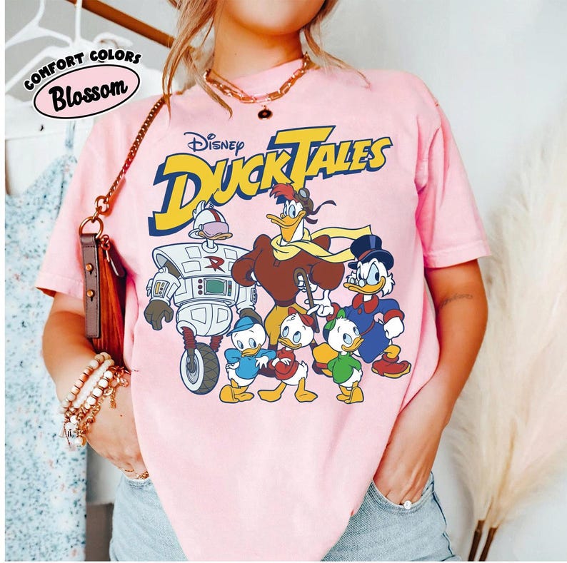 May include: Pink t-shirt featuring the Disney DuckTales logo and characters. The graphic includes Scrooge McDuck, Huey, Dewey, Louie, Launchpad McQuack, and Gizmoduck. The shirt also has the words "Comfort Colors" and "Blossom" printed on it.